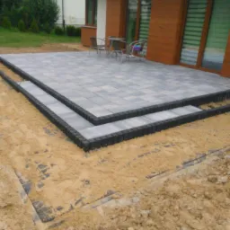 Nowo wyłożony taras z szarej kostki brukowej z czarnym obrzeżem, z dwoma stopniami, przed domem z brązową elewacją i zielonym trawnikiem.