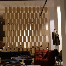 Nowoczesna aranżacja wnętrza z drewnianą ścianką działową z podwieszanych, prostokątnych element&oacute;w, czarną sofą sk&oacute;rzaną i fotelem w kolorze ceglastym oraz manekinem z marynarką.