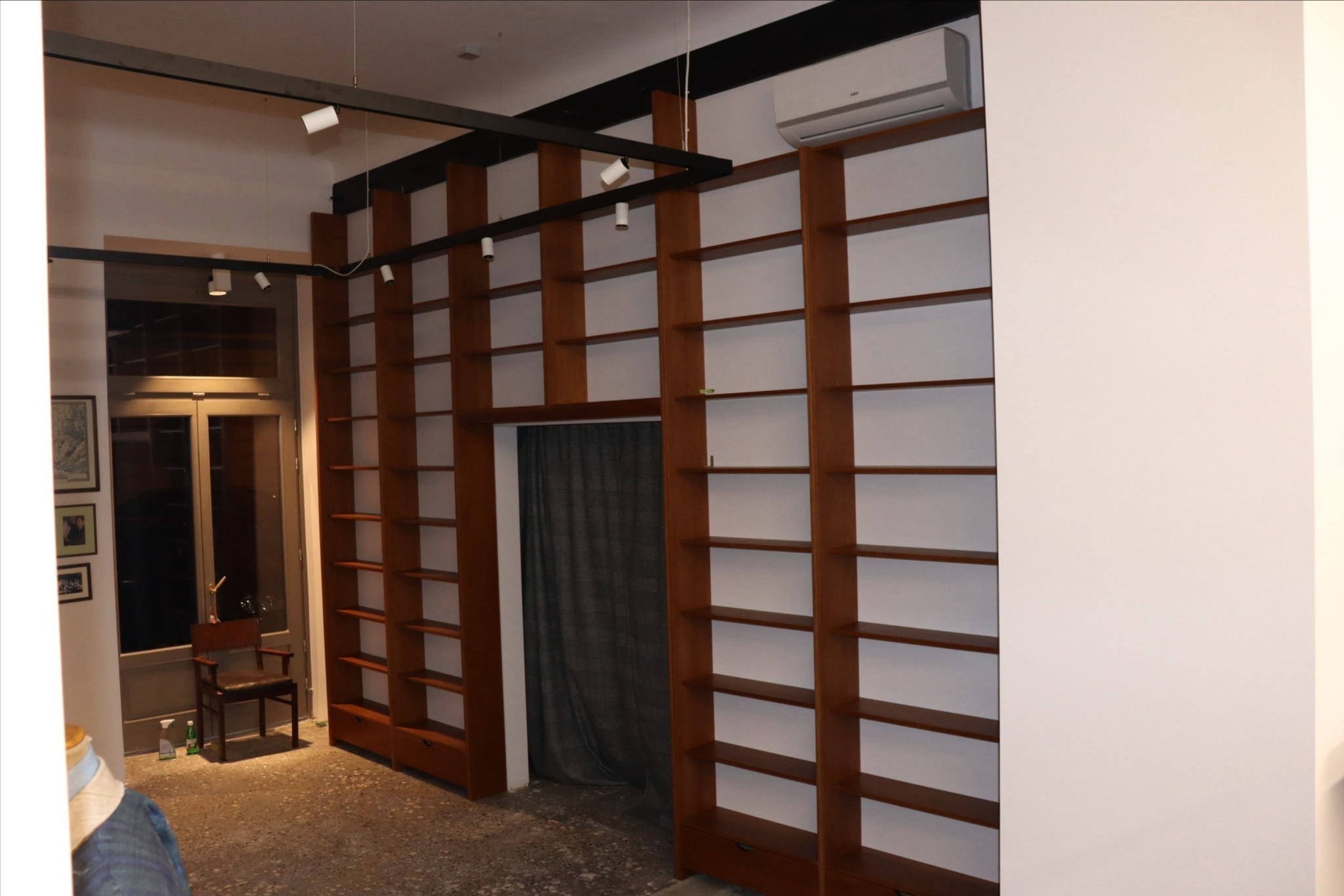 Wysokie, drewniane regały na książki, wbudowane od podłogi do sufitu, z oświetleniem punktowym na suficie, widok na drzwi z krzesłem w tle.