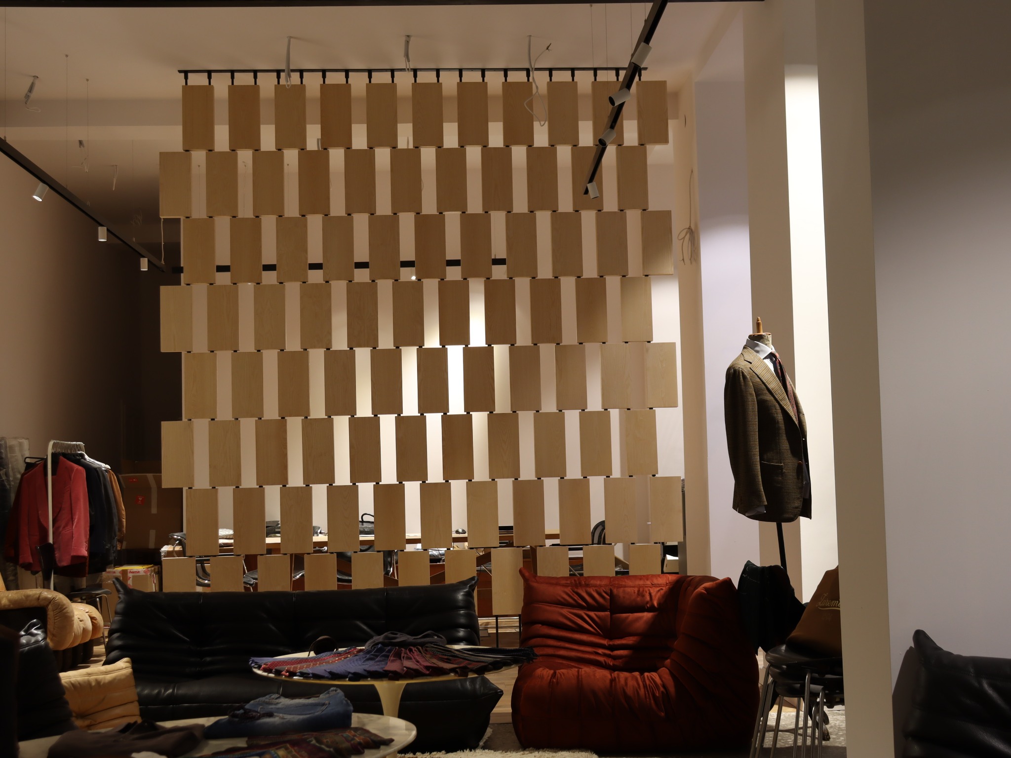 Nowoczesna aranżacja wnętrza z drewnianą ścianką działową z podwieszanych, prostokątnych elementów, czarną sofą skórzaną i fotelem w kolorze ceglastym oraz manekinem z marynarką.