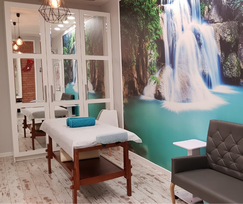 Jasne wnętrze gabinetu masażu z łóżkiem przykrytym białym prześcieradłem i turkusowym ręcznikiem, na ścianie fototapeta z wodospadem, obok szara sofa i szafa z lustrami.