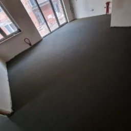 Świeżo wylana, gładka, ciemnoszara posadzka w surowym wnętrzu z widocznymi instalacjami na ścianach i oknami z widokiem na budowę.