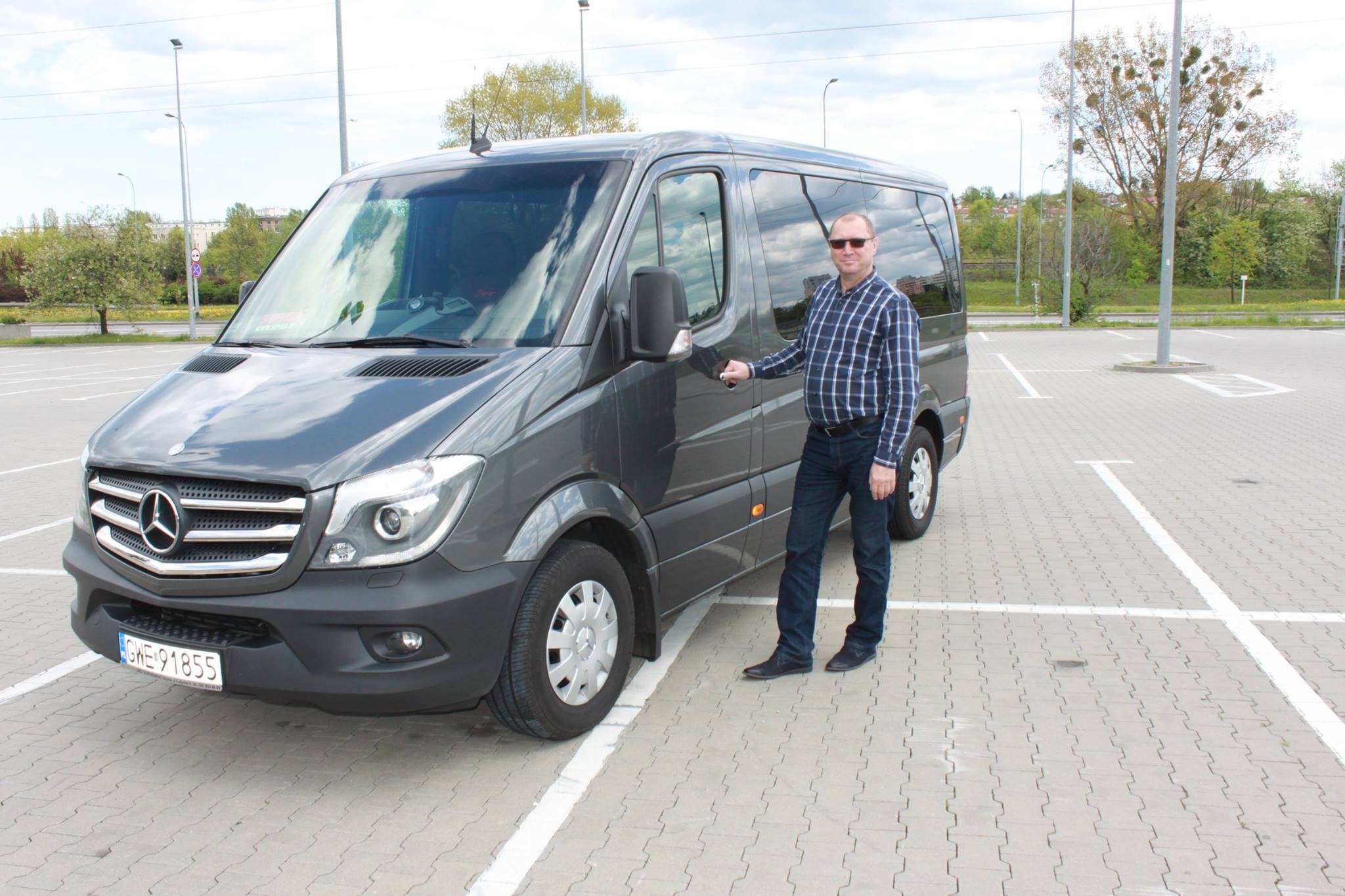 Szary bus Mercedes-Benz Sprinter z kierowcą w okularach i kraciastej koszuli na parkingu.