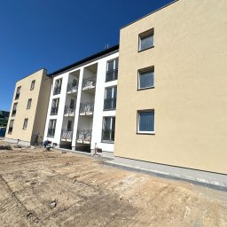 Nadzory budowlane JC Jacek Cząstkiewicz - Budowa nowego bloku mieszkalnego z beżową elewacją i białymi balkonami, widoczne prace ziemne i robotnicy przygotowujący teren.