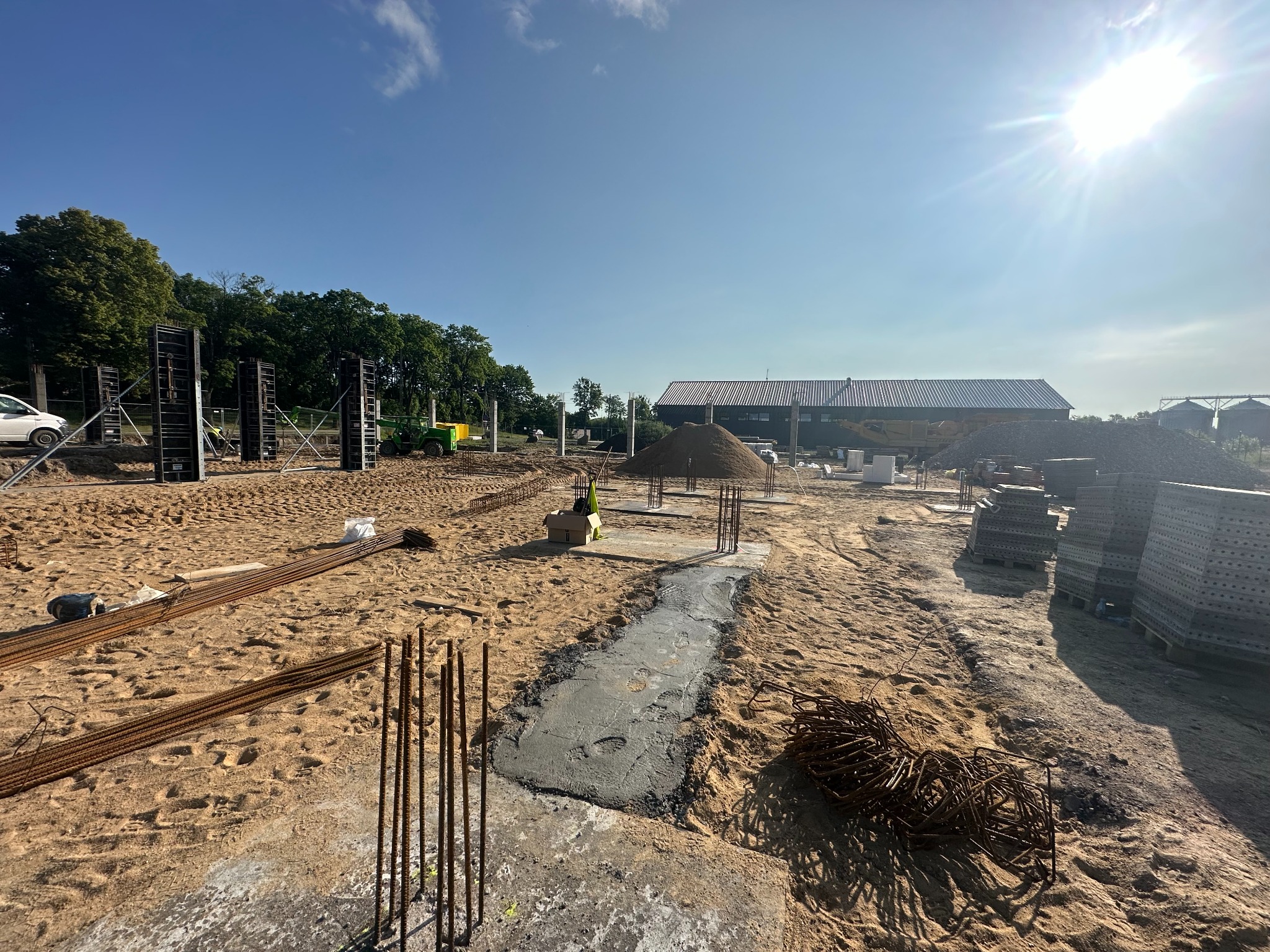 Fundamenty w trakcie wylewania, widoczne zbrojenia, szalunki i sterta piasku na placu budowy w słoneczny dzień.