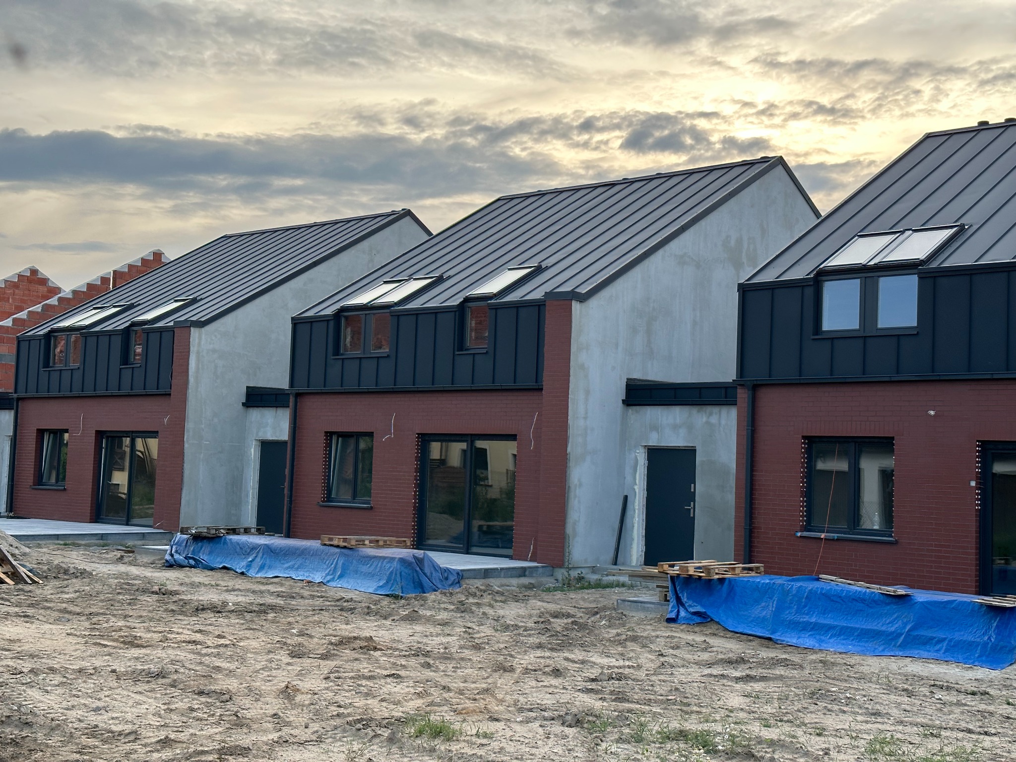 Nowe domy w zabudowie szeregowej z czerwonej cegły i szarego tynku, z ciemnymi dachami i oknami, częściowo przykryte plandekami na placu budowy.