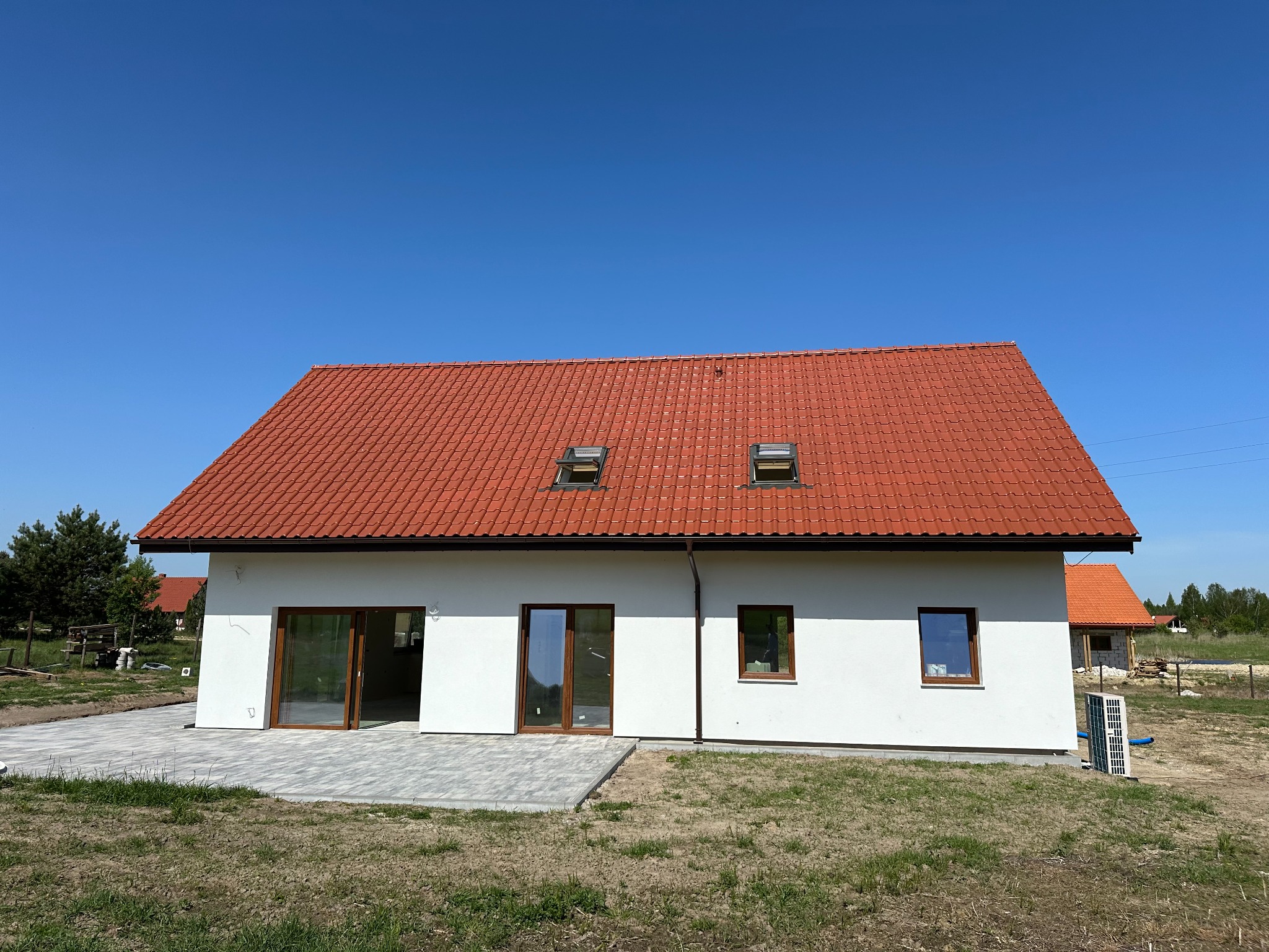 Nowo wybudowany, parterowy dom z czerwoną dachówką i dwoma oknami dachowymi, z szarą kostką brukową tworzącą taras, widoczny klimatyzator na zewnątrz.
