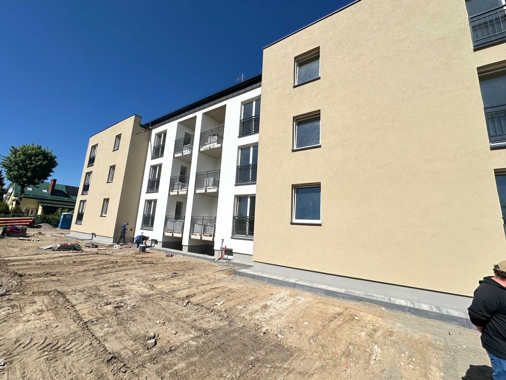 Budowa nowego bloku mieszkalnego z beżową elewacją i białymi balkonami, widoczne prace ziemne i robotnicy przygotowujący teren.