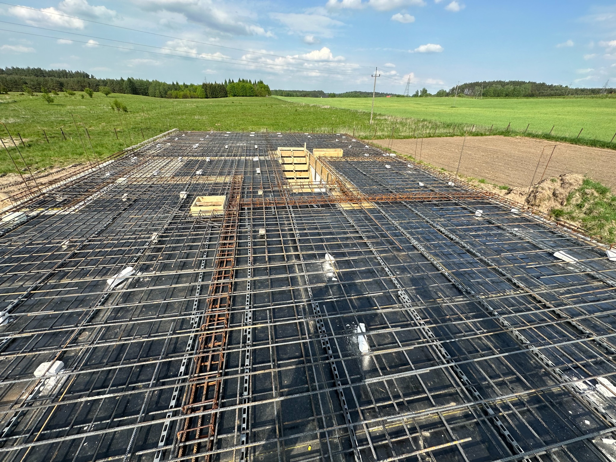 Zbrojenie fundamentów na czarnej hydroizolacji, widoczne drewniane szalunki i pręty wystające poza obrys, w tle zielone pola i las.