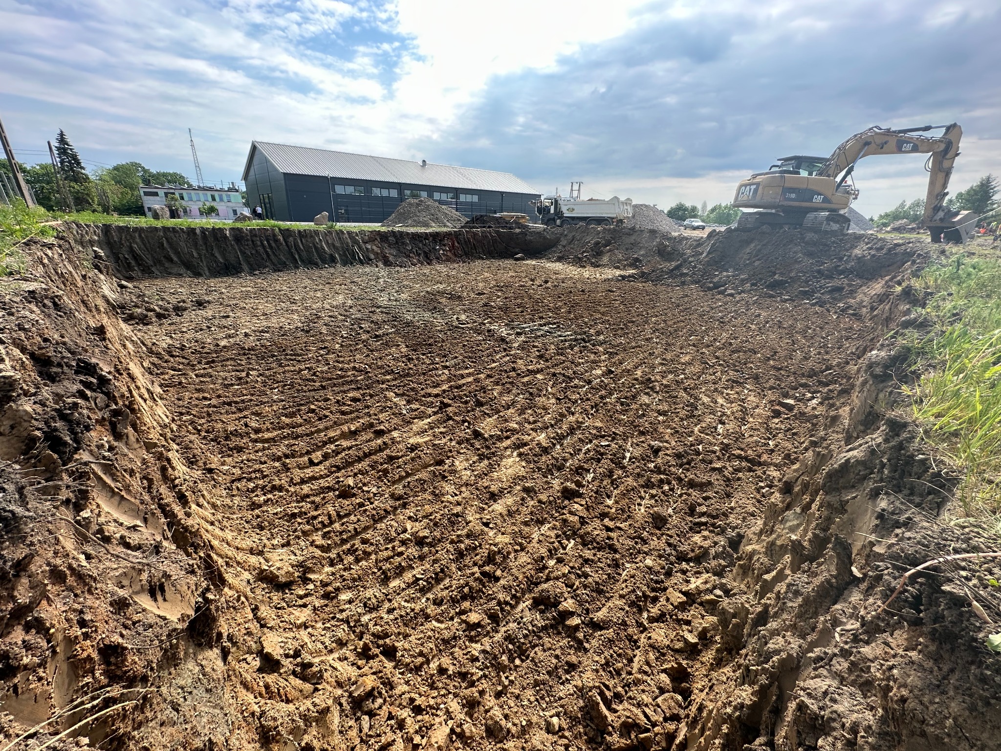 Wykop pod budowę z widocznymi śladami opon koparki, w tle budynek przemysłowy i ciężarówka z materiałami budowlanymi.