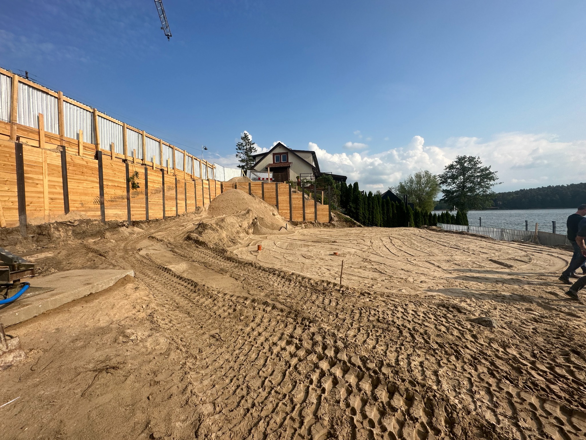 Teren budowy z przygotowanym podłożem, widoczny dom na wzniesieniu, drewniana palisada i częściowo metalowe ogrodzenie, oraz ślady opon ciężkiego sprzętu.