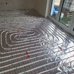 Białe rury systemu grzewczego ułożone spiralnie na czerwonej siatce, pokryte folią odblaskową w surowym wnętrzu, widoczne okno z elementami budowlanymi na zewnątrz.