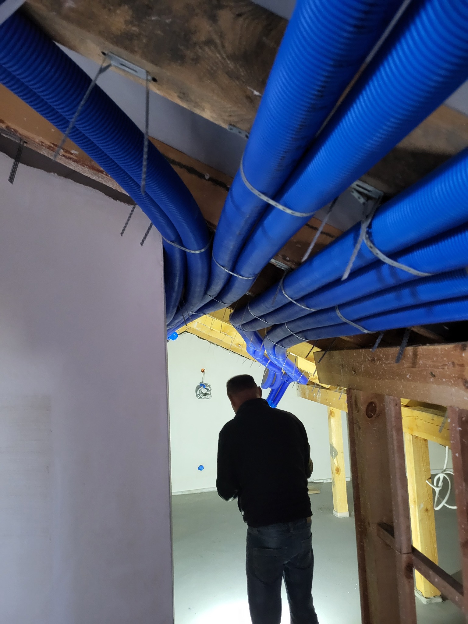 Niebieskie, karbowane rury wentylacyjne zamocowane pod drewnianym stropem na metalowych uchwytach, widoczne fragmenty konstrukcji szkieletowej budynku i sylwetka instalatora w ciemnym ubraniu.