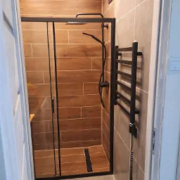 Nowoczesna kabina prysznicowa z czarną armaturą i grzejnikiem drabinkowym, brązowe płytki imitujące drewno na ścianach kabiny, szara podłoga, widok z zewnątrz kabiny.