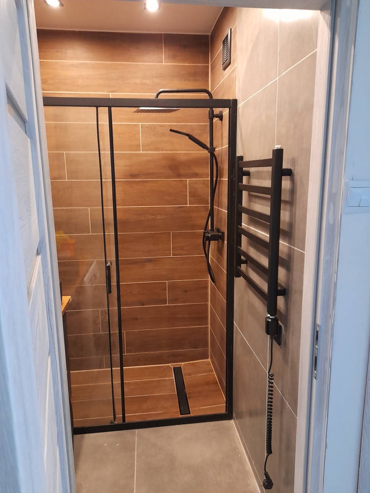 Nowoczesna kabina prysznicowa z czarną armaturą i grzejnikiem drabinkowym, brązowe płytki imitujące drewno na ścianach kabiny, szara podłoga, widok z zewnątrz kabiny.