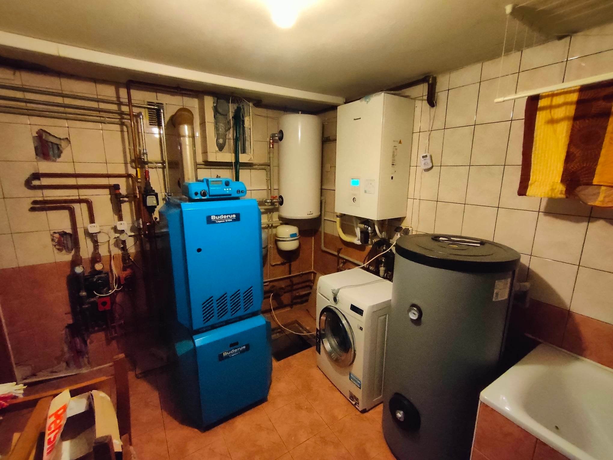 Kotłownia w piwnicy z niebieskim piecem Buderus, białym bojlerem, pralką i szarym zbiornikiem akumulacyjnym, widoczne rury i płytki ceramiczne na ścianach.