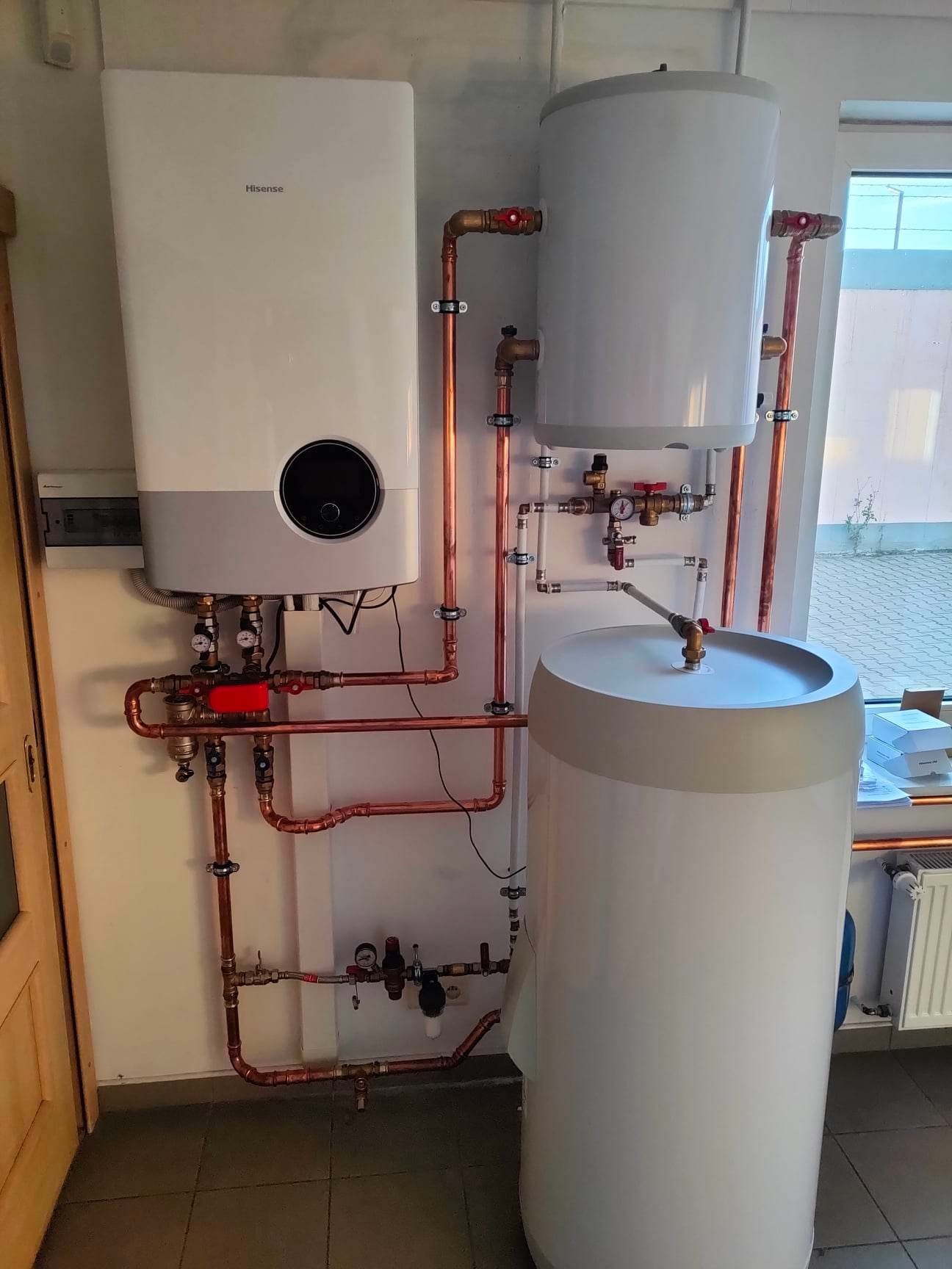 Nowoczesny piec grzewczy Hisense z dwoma zasobnikami ciepłej wody, połączony miedzianymi rurami, zainstalowany w pomieszczeniu z grzejnikiem i oknem.