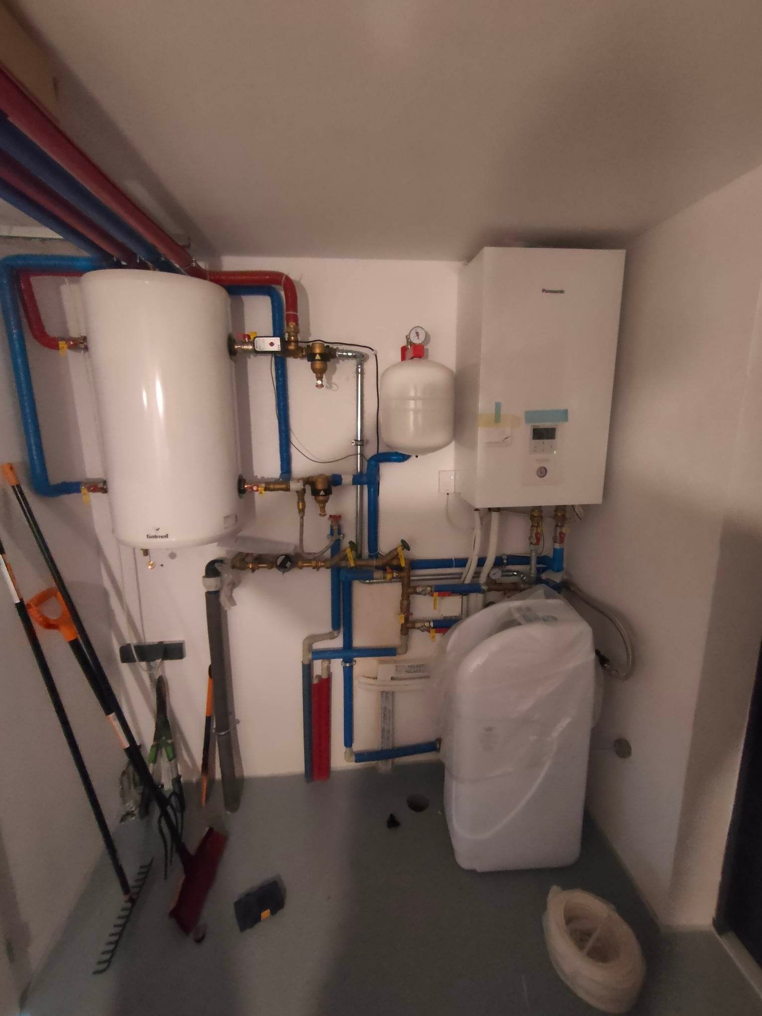Biały bojler, piec gazowy i zmiękczacz wody w folii, połączone instalacją z rur miedzianych i PCV w małym pomieszczeniu gospodarczym z narzędziami ogrodniczymi opartymi o ścianę.