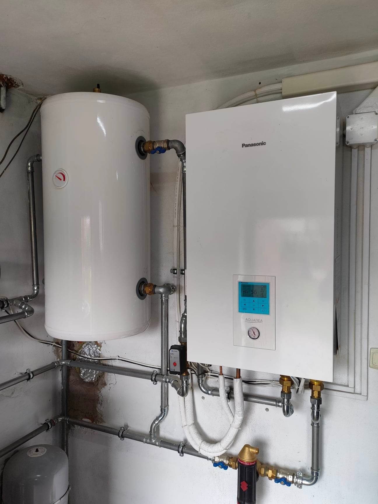Biały piec Panasonic Aquarea z wyświetlaczem i zbiornik na wodę, połączone rurami w kotłowni, widoczny filtr i izolacja na rurach.