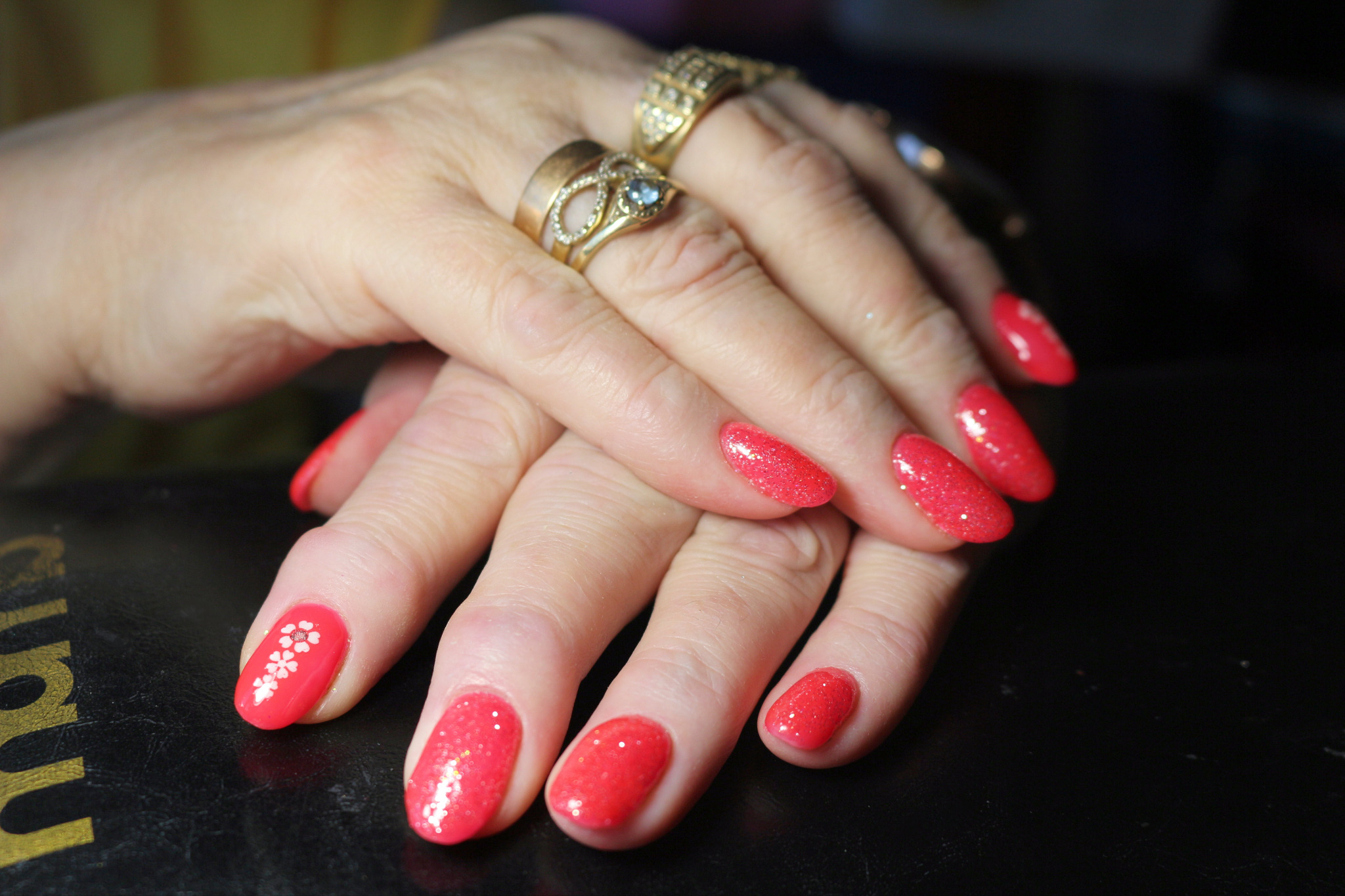 Dłonie kobiety z czerwonym, błyszczącym manicure hybrydowym, na jednym paznokciu wzór z białych kwiatków, ozdobione złotymi pierścionkami.