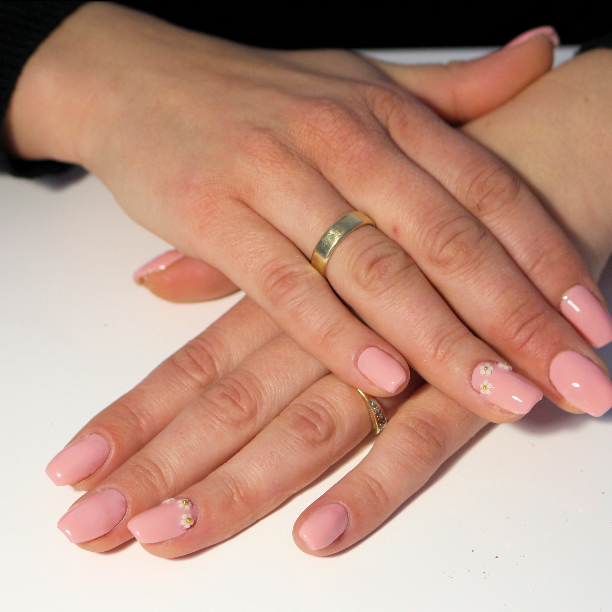 Dłonie kobiety z delikatnym, różowym manicure hybrydowym i kwiatowymi zdobieniami na dwóch paznokciach, na jednym palcu widoczny złoty pierścionek.