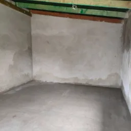 Surowe wnętrze pokoju w trakcie prac wykończeniowych: ściany pokryte świeżym tynkiem, widoczne drewniane elementy konstrukcyjne stropu i podłoga wylewka betonowa.