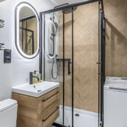 Nowoczesna łazienka z kabiną prysznicową w czarnej ramie, umywalką z szafką z jasnego drewna, podświetlanym lustrem i ścianą z wzorem jodełki.