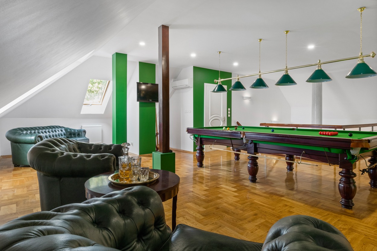 Stylowe poddasze z bilardem, zielone akcenty na ścianach, skórzane sofy typu Chesterfield, parkiet w jodełkę i kryształowa karafka z whisky na stoliku.