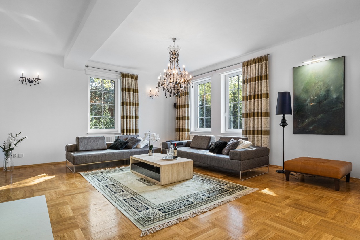 Elegancki salon z parkietem w jodełkę, dwoma szarymi sofami, kryształowym żyrandolem i obrazem z podświetleniem, uzupełniony beżowym dywanem i drewnianym stolikiem kawowym z winem.