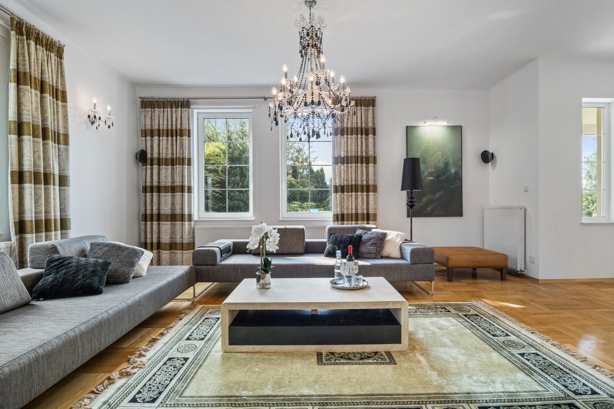 Jasne wnętrze salonu z szarymi sofami, drewnianą podłogą w jodełkę, wzorzystym dywanem, kryształowym żyrandolem i obrazem na ścianie.