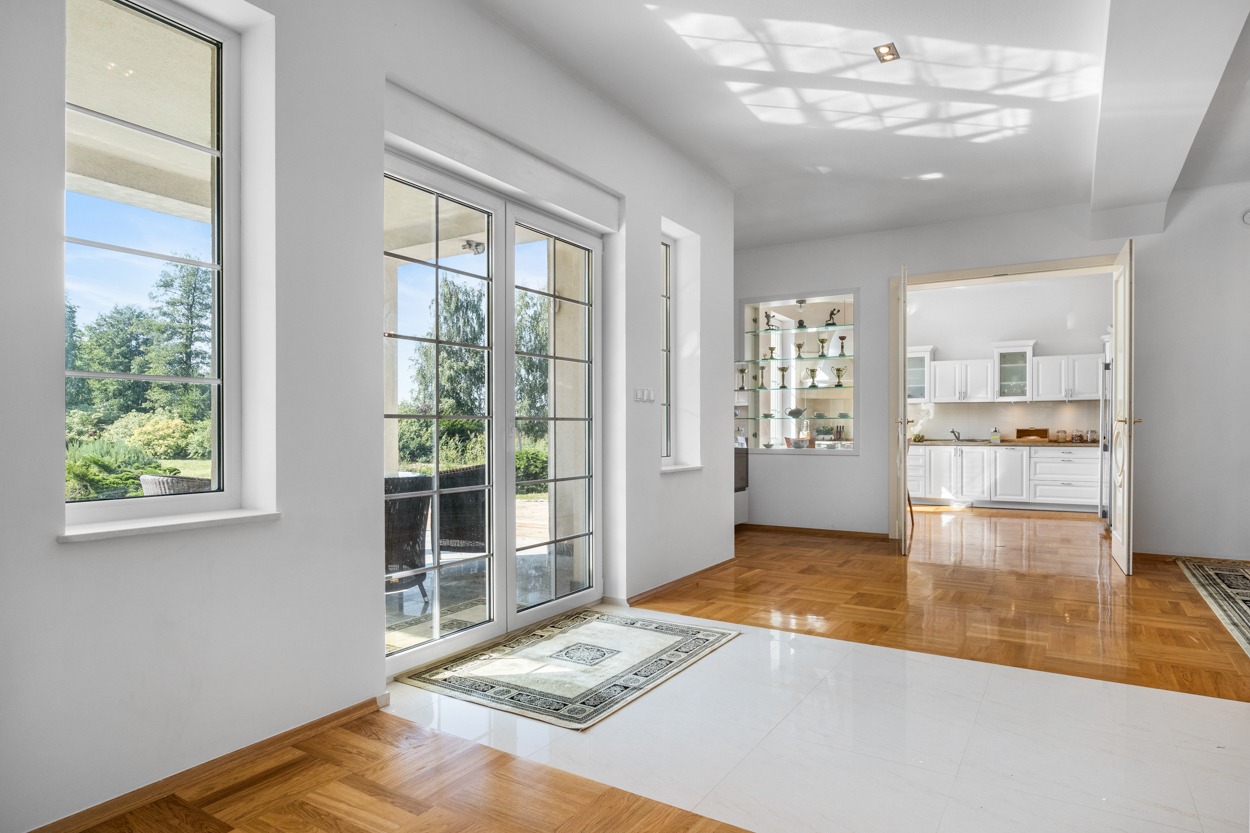 Jasne wnętrze domu z dużymi oknami wychodzącymi na ogród, widoczna biała kuchnia w tle i drewniana podłoga przechodząca w białe płytki.
