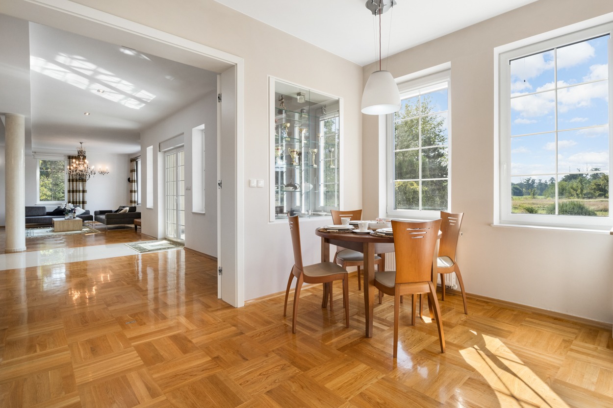 Jasne wnętrze domu z parkietem w jodełkę, okrągłym stołem jadalnym z nakryciem i widokiem na salon z żyrandolem kryształowym.