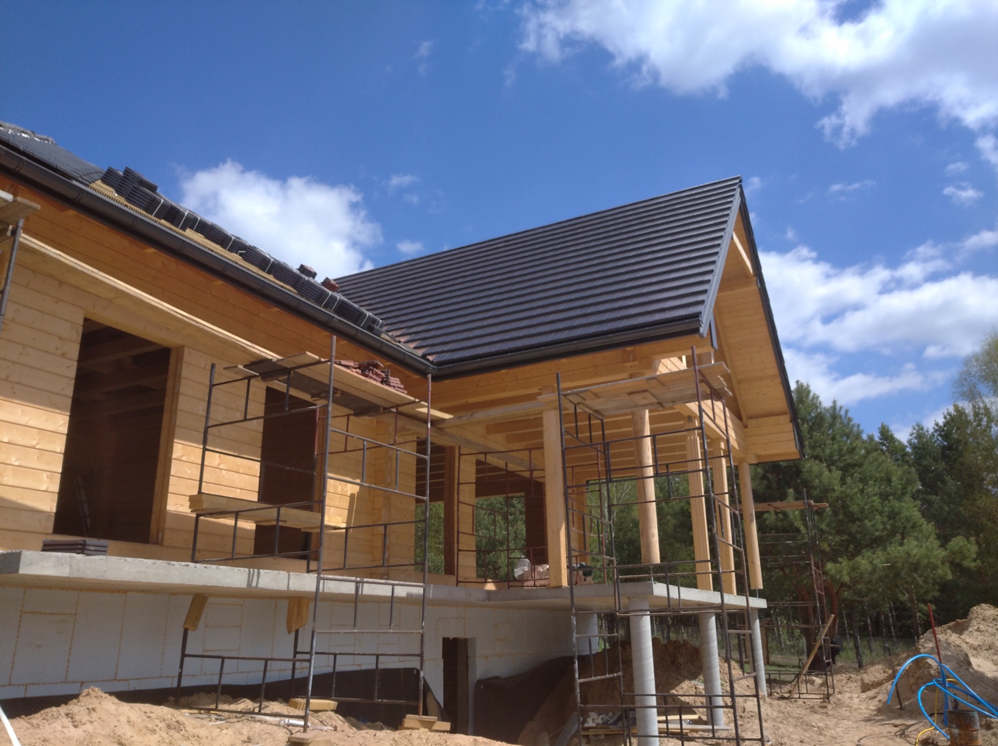 Budowa domu w technologii szkieletowej: widok z dołu na drewnianą konstrukcję, rusztowania i częściowo położony ciemny dach na tle błękitnego nieba z chmurami.