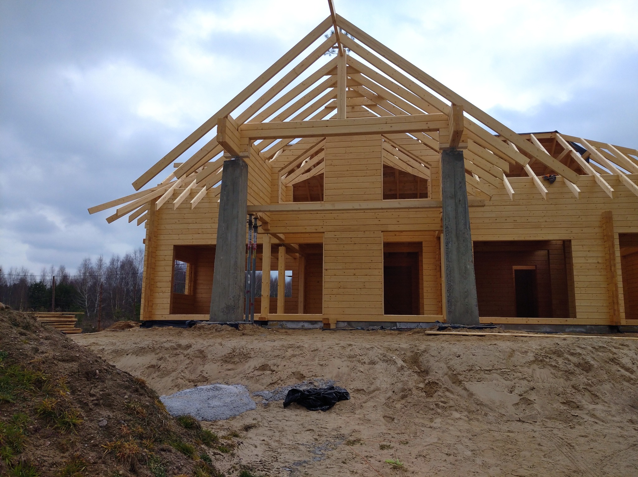 Surowy stan budowy domu z bali, widoczne betonowe filary, drewniana konstrukcja dachu i ściany z bali, teren budowy z piaskiem i stertą desek w tle.
