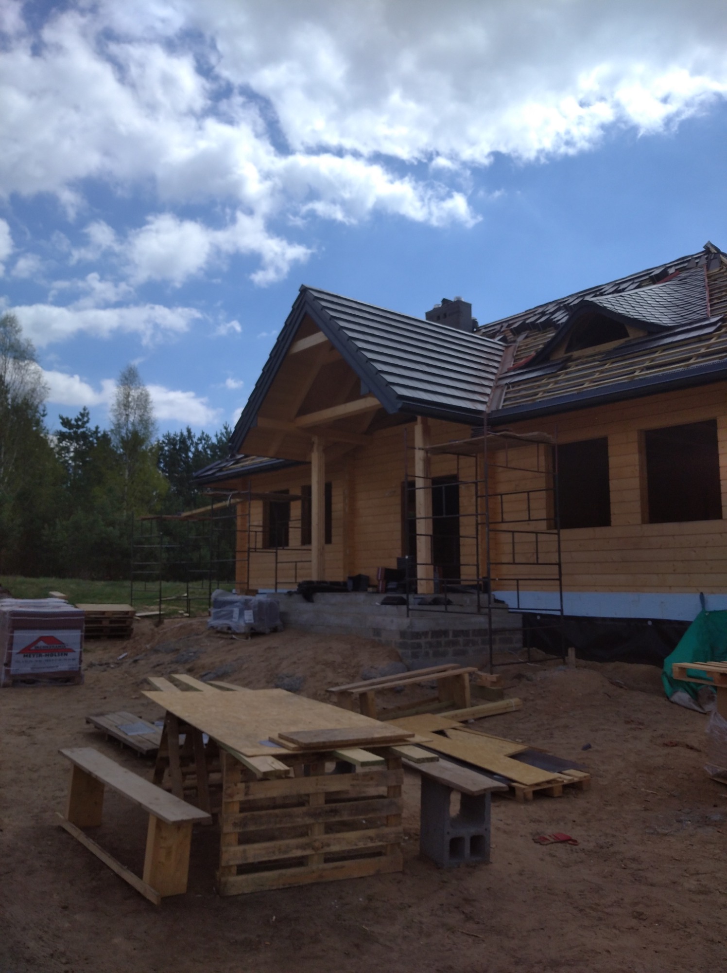 Budowa domu z bali w trakcie realizacji: widoczne rusztowania, częściowo położony dach z czarnej dachówki, palety z materiałami budowlanymi i prowizoryczny stół z palet na placu budowy.