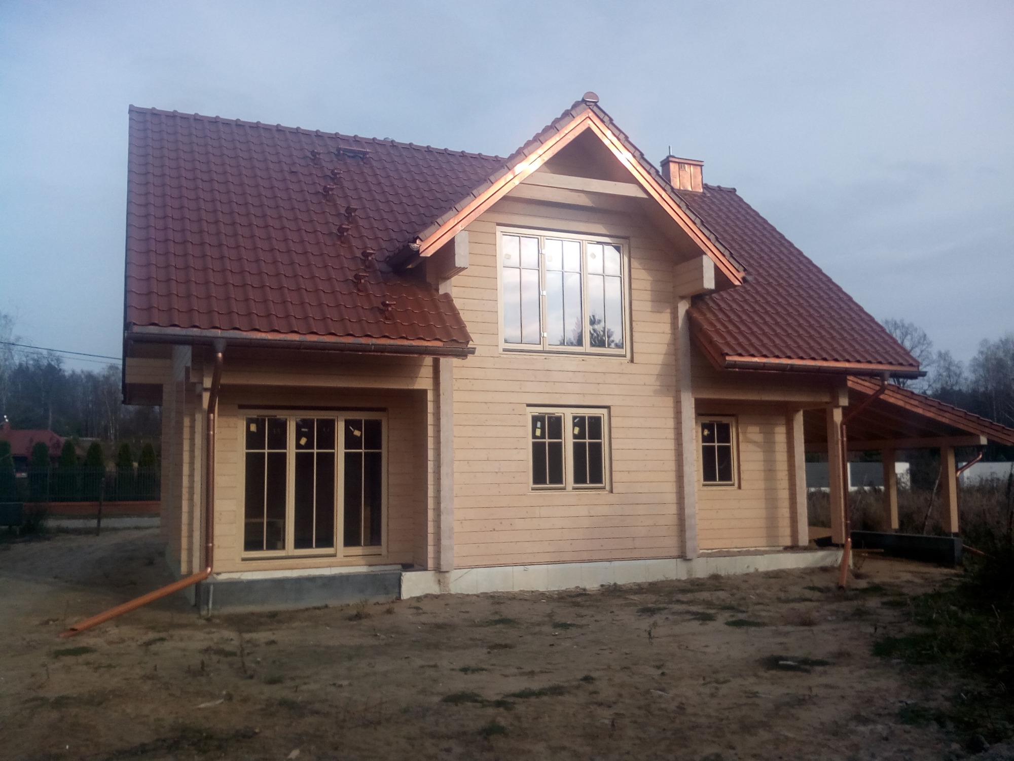 Surowy, dwukondygnacyjny budynek z bali z brązowym dachem z blachodachówki i miedzianymi rynnami, widoczny w pochmurny dzień.