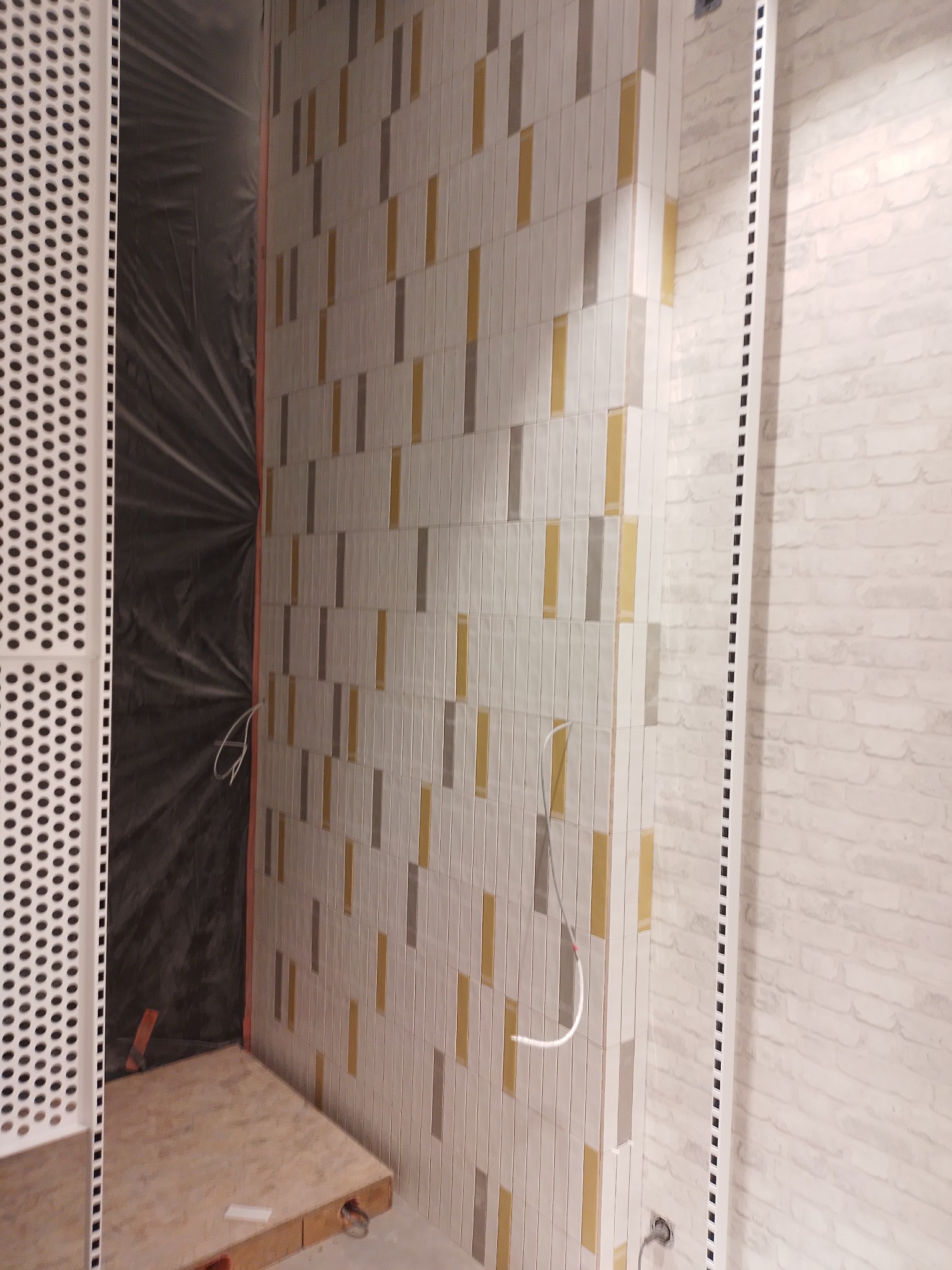 Fragment łazienki w trakcie remontu: ułożone płytki w pionowe paski w odcieniach szarości i złota na ścianie, zabezpieczona folią strefa prysznicowa, widoczne przewody elektryczne i elementy...