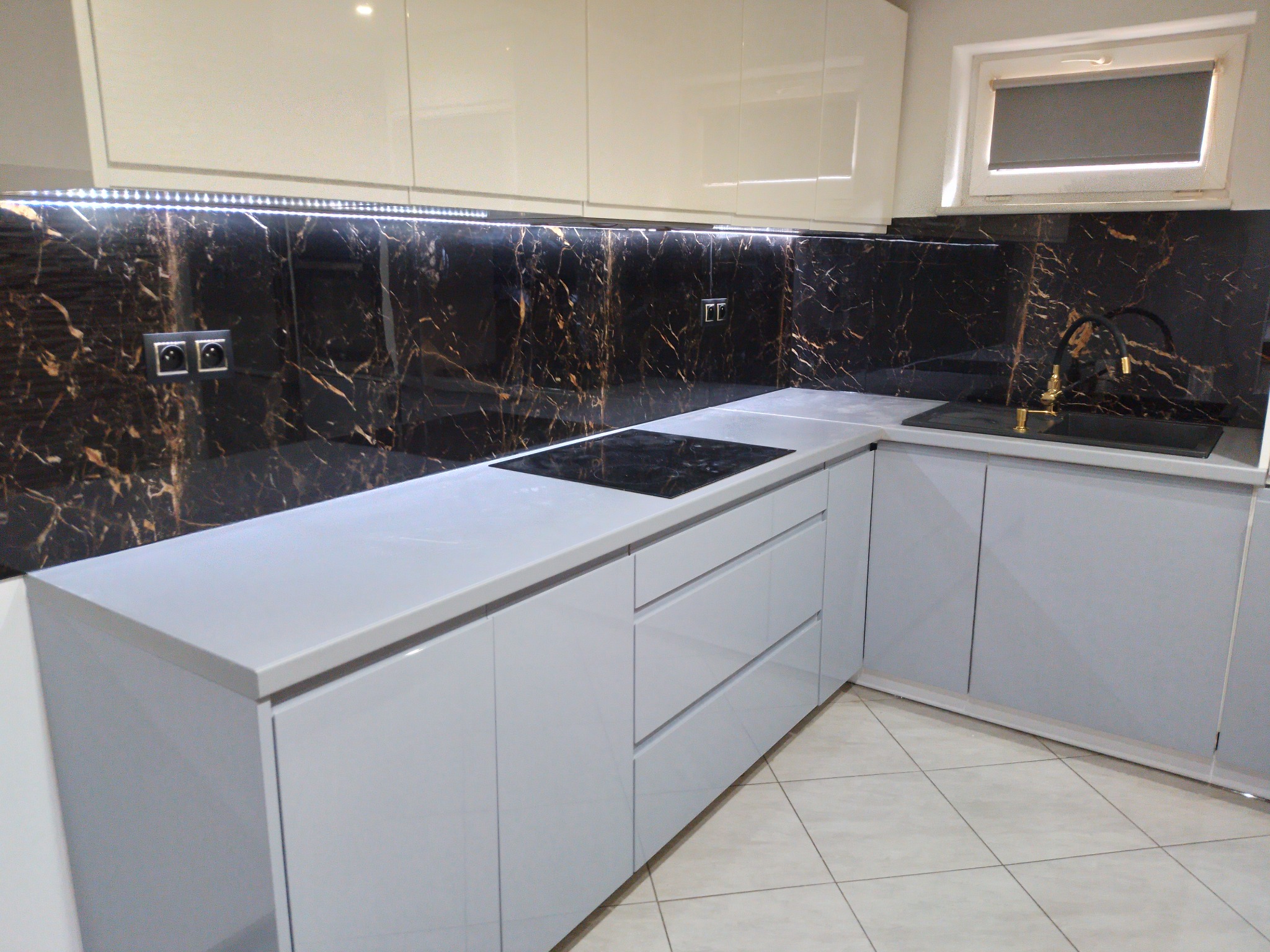 Nowoczesna kuchnia z szarymi, lakierowanymi szafkami, blatem z jasnego konglomeratu i czarnym panelem szklanym z marmurkowym wzorem, oświetlonym listwą LED.