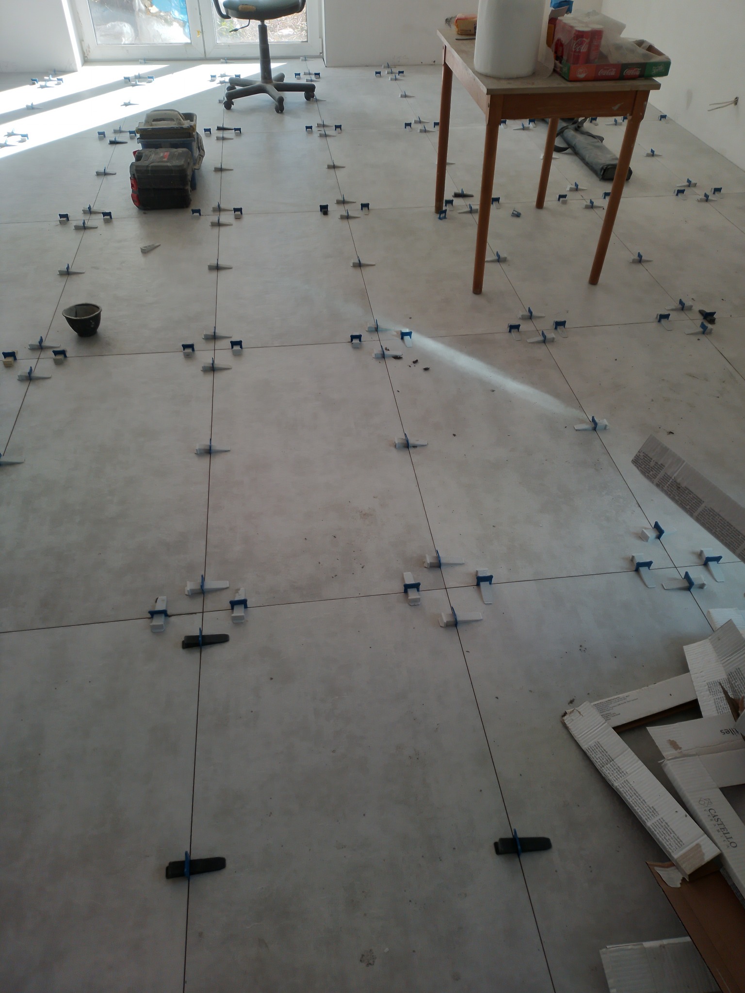 Podłoga w trakcie układania płytek, widoczne krzyżyki dystansowe, poziomica, wiadro, narzędzia w skrzynce, kartony po płytkach i stół z materiałami budowlanymi.