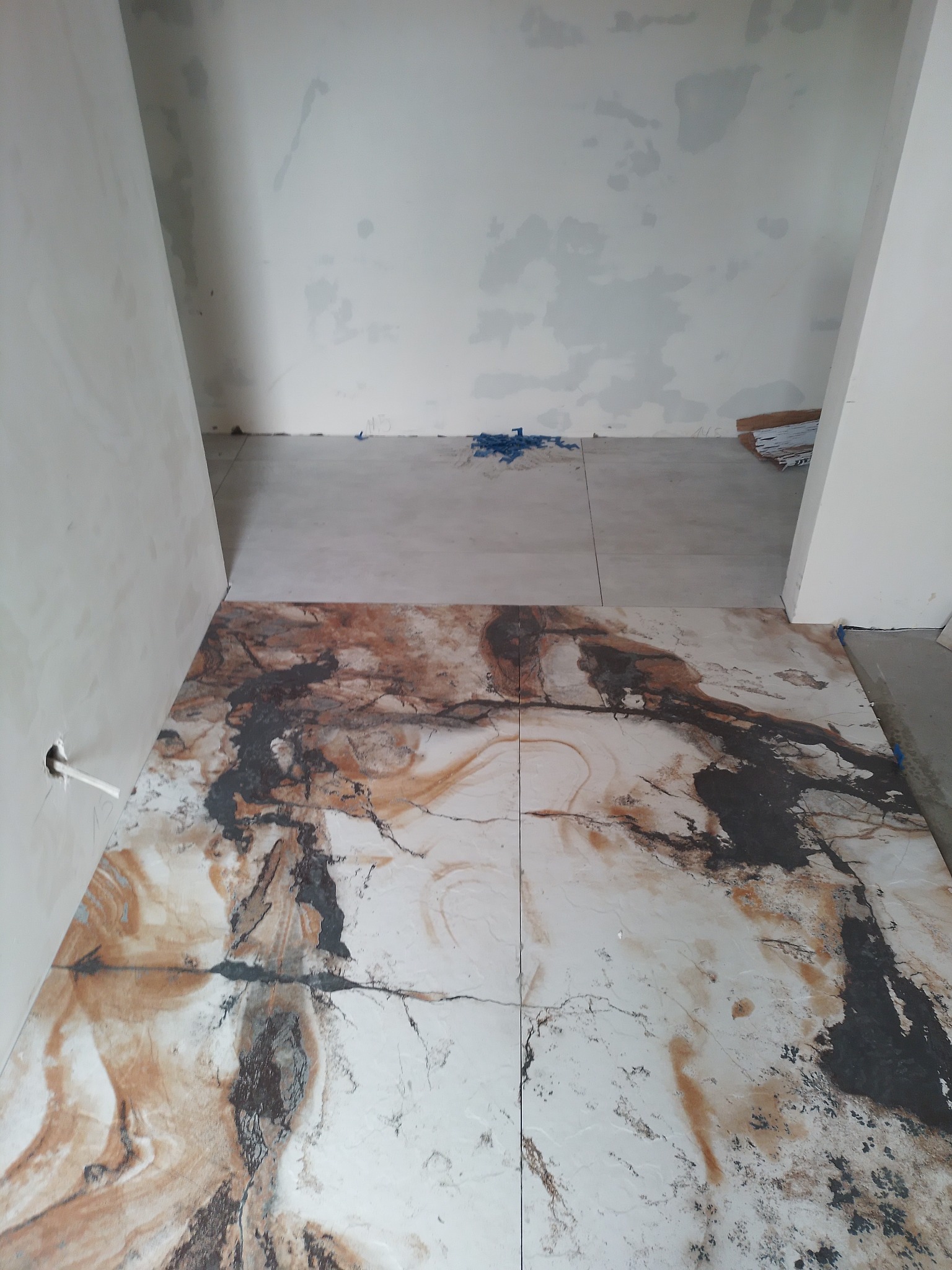 Fragment podłogi z ułożonymi płytkami imitującymi marmur w odcieniach brązu i szarości, widoczne niedokończone ściany i fragmenty podłoża przygotowanego pod płytki.