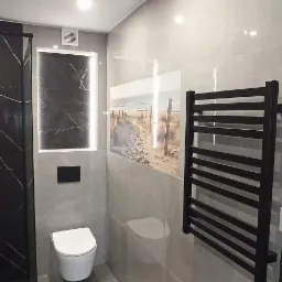 Nowoczesna łazienka z szarą glazurą, podwieszaną toaletą, czarnym grzejnikiem drabinkowym i dekoracyjnym panelem ściennym z motywem plaży.