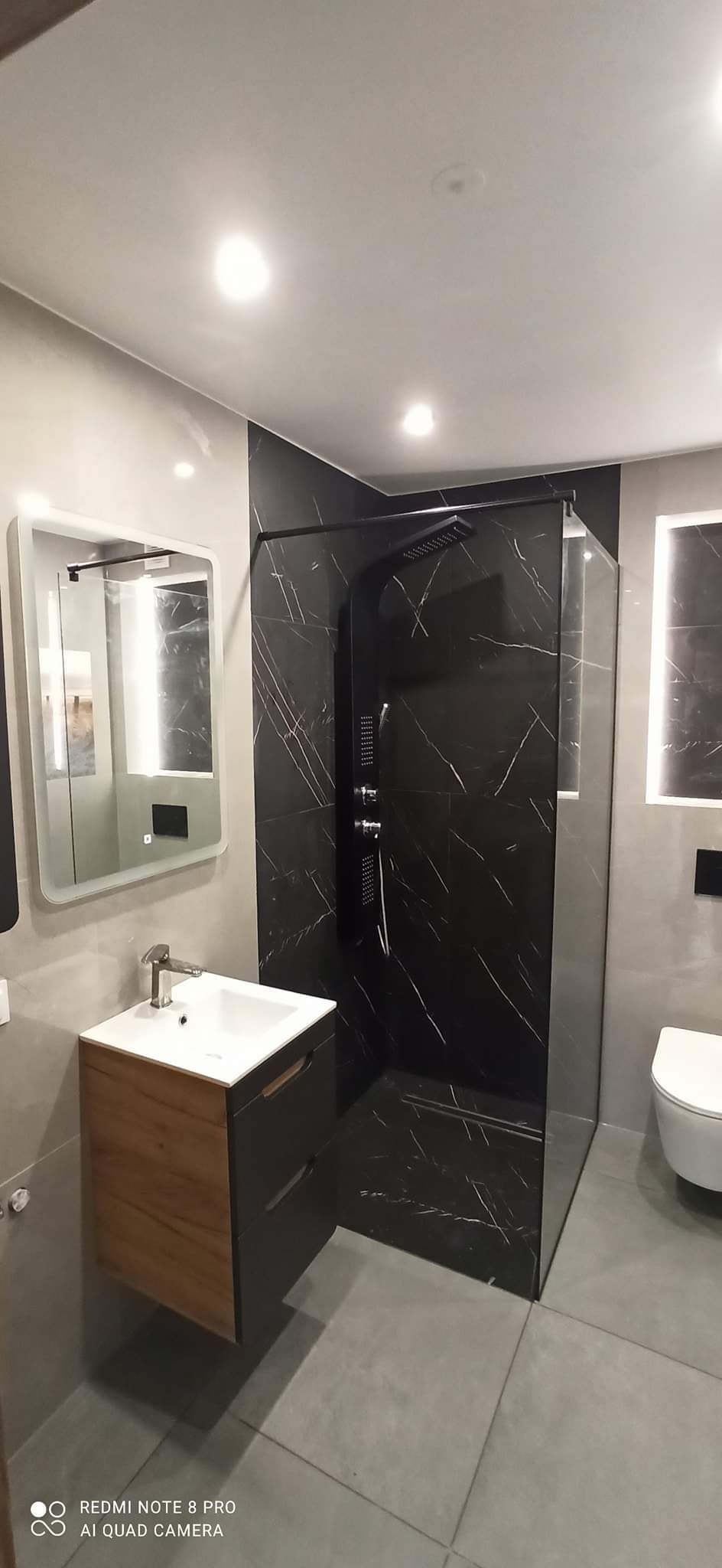 Nowoczesna łazienka z kabiną prysznicową wyłożoną czarnym marmurem, umywalką z szafką w kolorze drewna i szarą płytką podłogową.