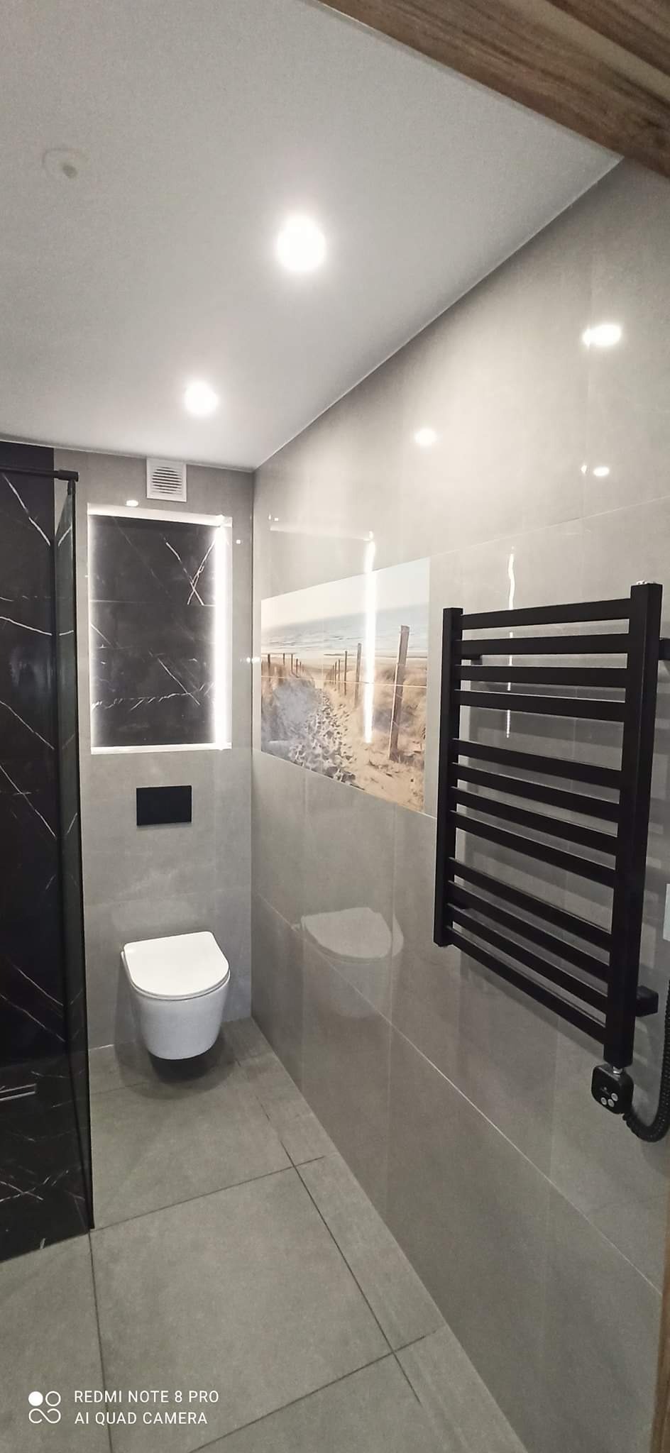 Nowoczesna łazienka z szarą glazurą, podwieszaną toaletą, czarnym grzejnikiem drabinkowym i dekoracyjnym panelem ściennym z motywem plaży.