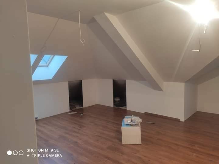 Wykończone poddasze z drewnianą podłogą, białymi ścianami i oknem dachowym. W głębi widoczne dwa otwory w ścianie. Na podłodze leżą resztki materiałów budowlanych.