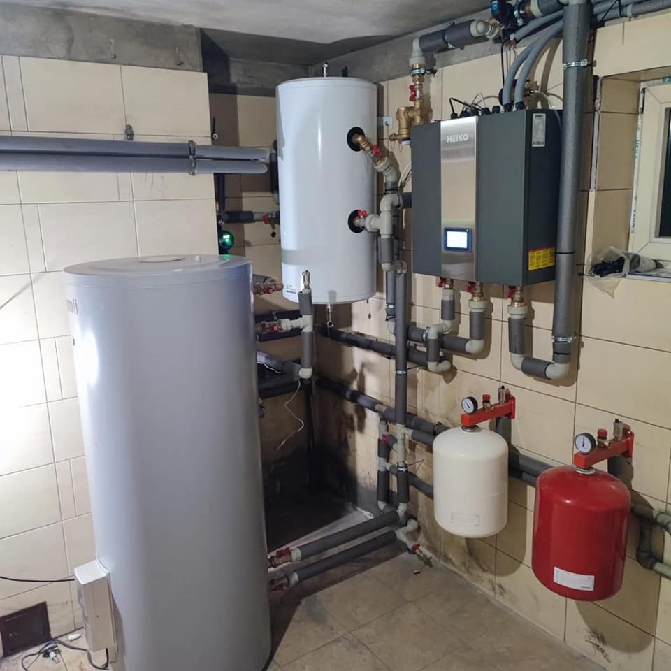 Złożona instalacja grzewcza z dwoma zbiornikami akumulacyjnymi, pompą ciepła Heiko i licznymi rurami w piwnicy z beżowymi płytkami ściennymi.