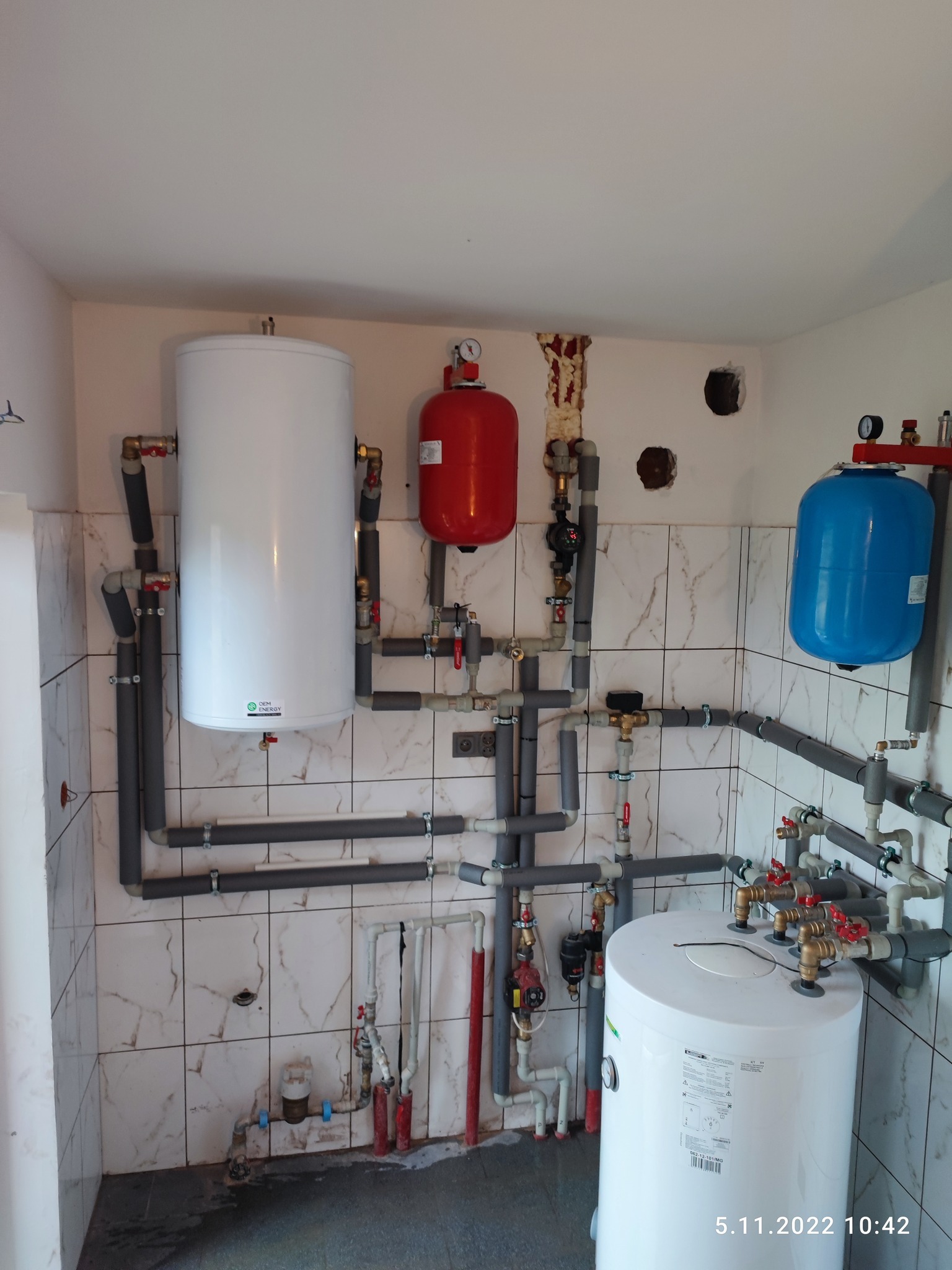 Kompletna instalacja grzewcza z trzema zbiornikami (białym, czerwonym i niebieskim) oraz szarymi rurami, zamontowana na ścianie wyłożonej marmurkowymi płytkami ceramicznymi.