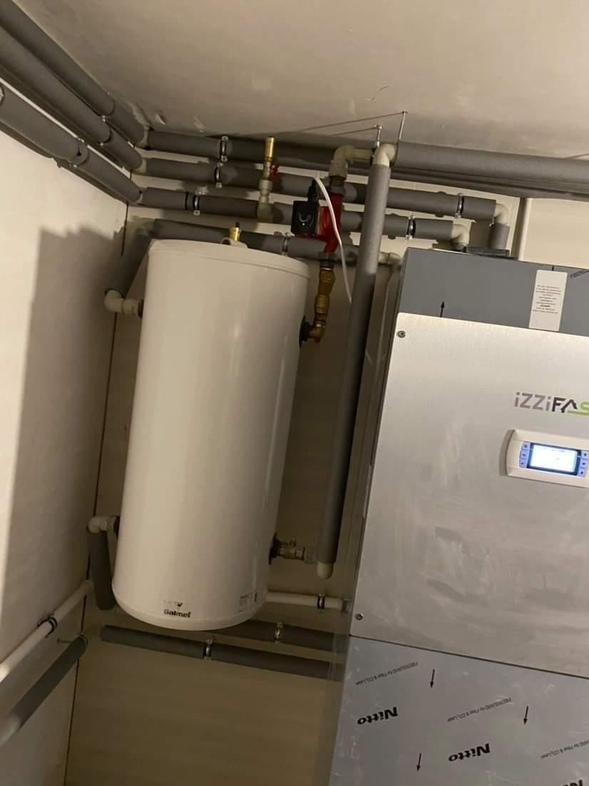 Biały bojler Galmet zawieszony na ścianie, z licznymi rurami w izolacji termicznej, podłączony do urządzenia grzewczego IZZIFAS z wyświetlaczem.