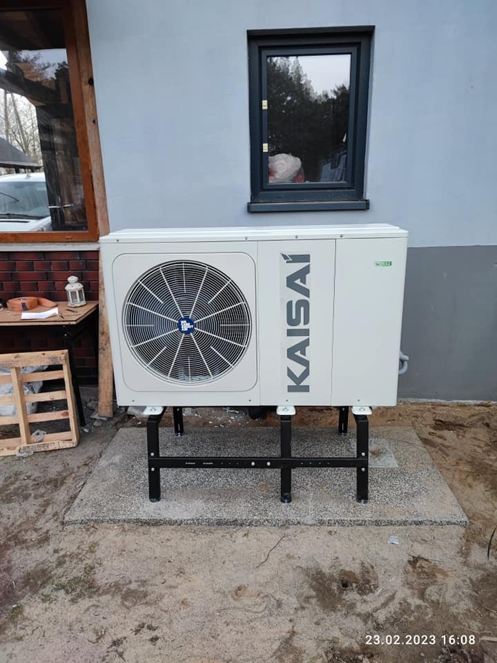 Zewnętrzna jednostka pompy ciepła Kaisai zamontowana na czarnej, stalowej konstrukcji na betonowej podstawie, z widocznym fragmentem elewacji budynku i oknami.