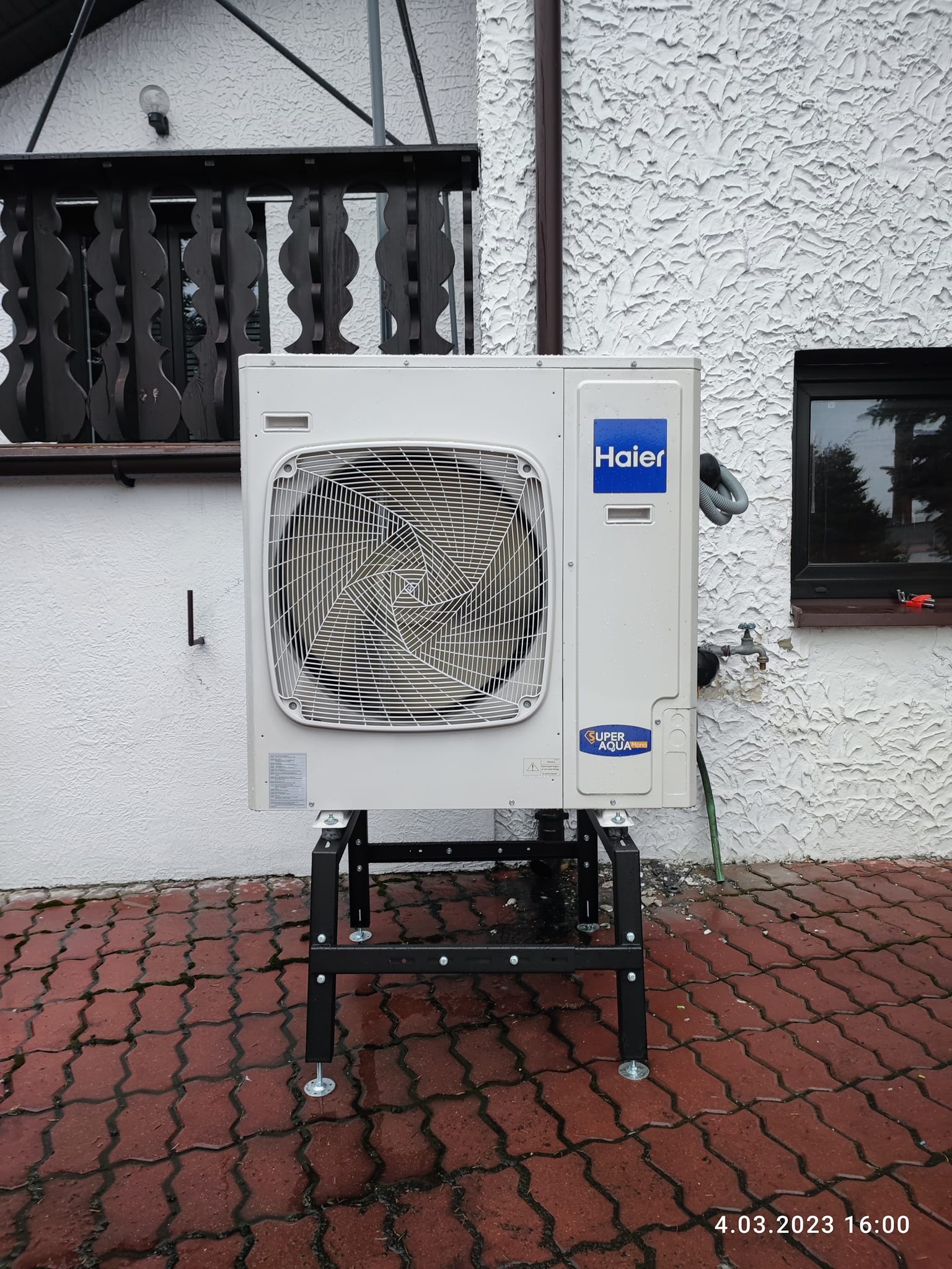 Biała pompa ciepła Haier Super Aqua na czarnym metalowym stojaku, zamontowana przy białej, chropowatej ścianie budynku z widocznym oknem i fragmentem balkonu z drewnianą balustradą.