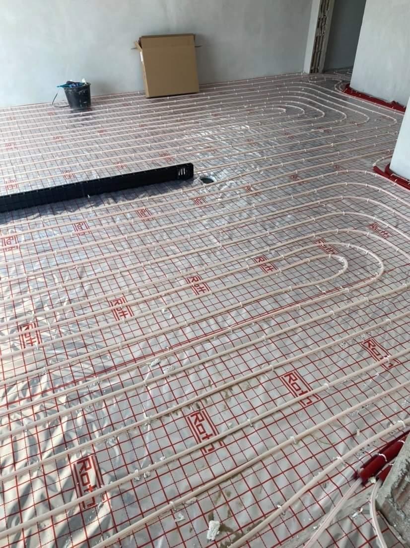 Rozłożone rury ogrzewania podłogowego na folii z siatką, widoczne narzędzia i przygotowane otwory w podłodze, w tle karton i wiadro.